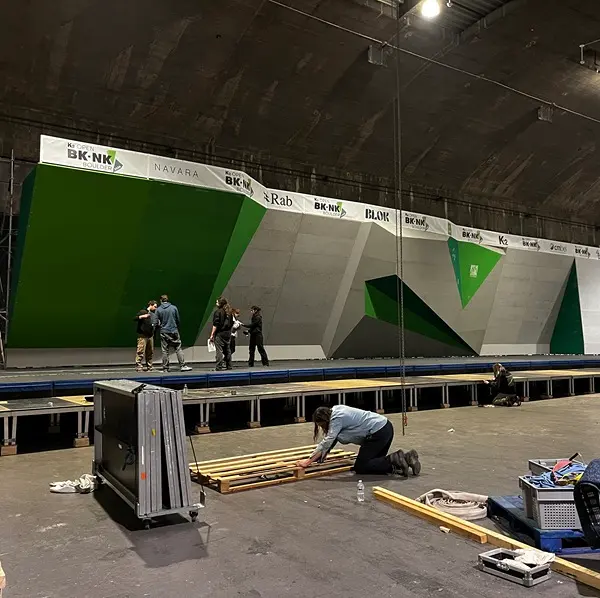 Er wordt binnen een klimwand voor een wedstrijd opgebouwd. Een aantal mensen staat voor grote, geometrische groene en grijze klimpanelen, terwijl anderen op de vloer op de voorgrond met gereedschap en materialen aan het werk zijn.